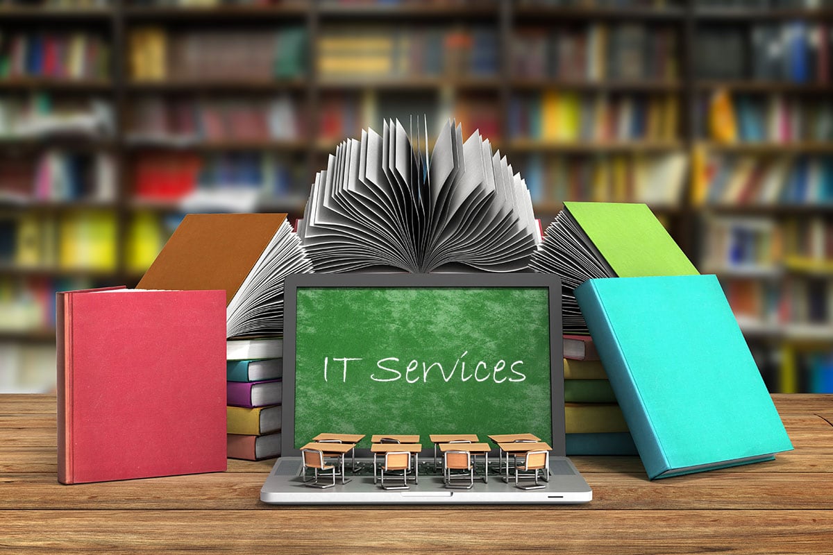 In a room full of books, a laptop shows a green chalkboard with the text "IT Services" written on it.