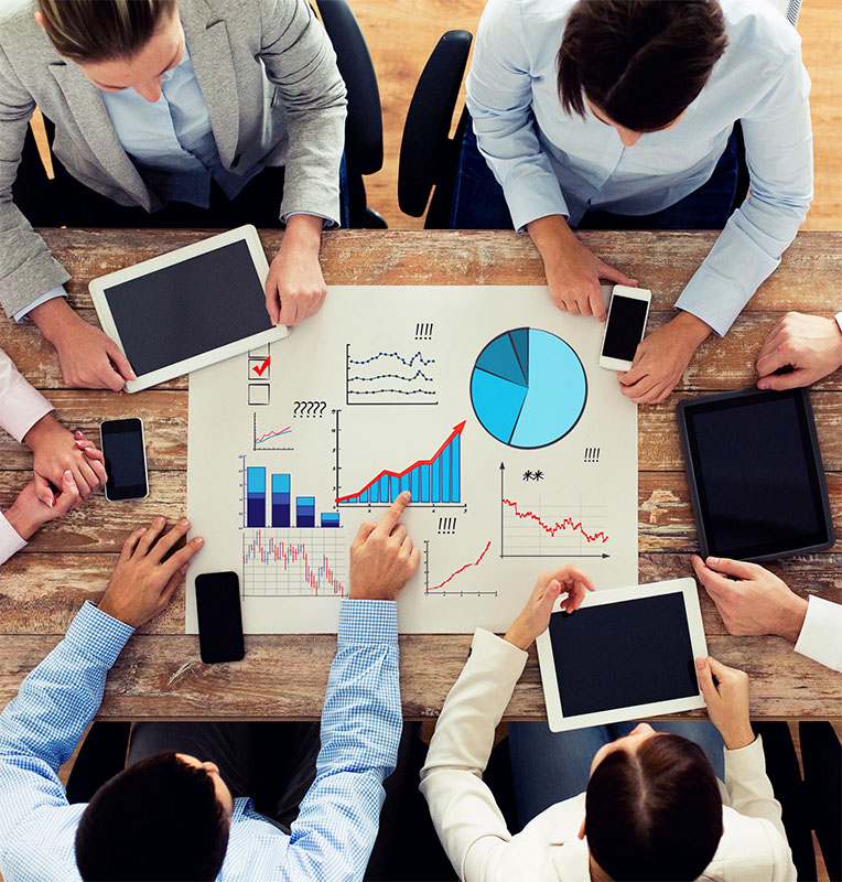 A group of people is gathered around a table covered with charts and graphs, analyzing data displayed on paper and digital devices, such as tablets and smartphones. The individuals are gesturing towards the visual data, discussing insights collaboratively.